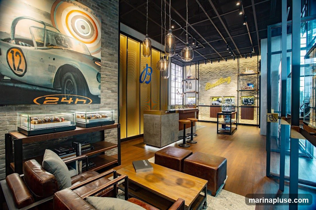The interior of a Breitling watch store features a modern and luxurious design. A large mural of a vintage race car adorns one wall, while display cases showcase various watches. A central counter with stools and comfortable leather seating areas create a welcoming atmosphere. The brand's logo is prominently displayed, and shelves are stocked with watches and accessories.