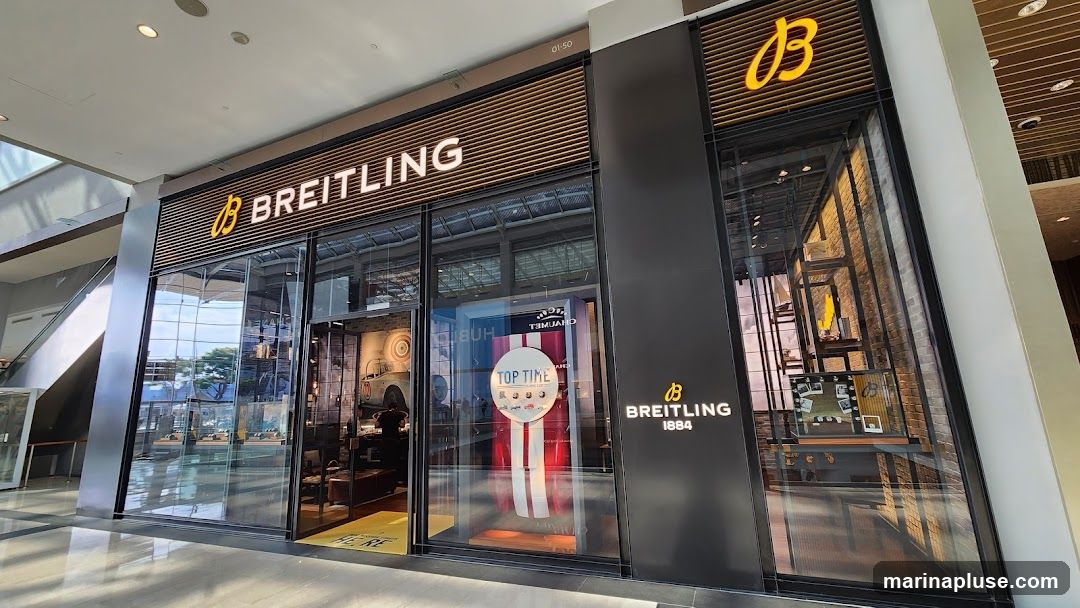 The exterior of a Breitling watch store. The store has large glass windows and doors, with the Breitling logo and name prominently displayed above the entrance. Inside, watches and displays are visible. A promotional display for "Top Time" watches is featured in the window.