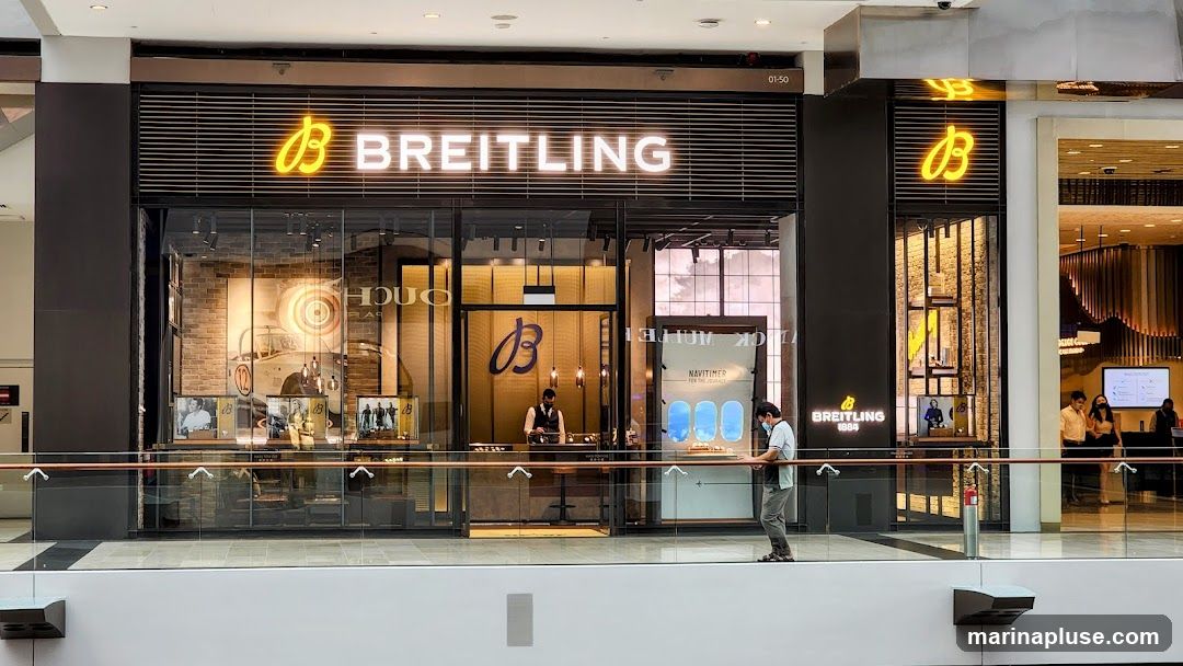The exterior of a Breitling watch store. The store has a large glass facade with the Breitling logo and name prominently displayed in neon lights above the entrance. Inside, watches are displayed in illuminated cases. A staff member stands behind a counter, and a customer is looking at a display.