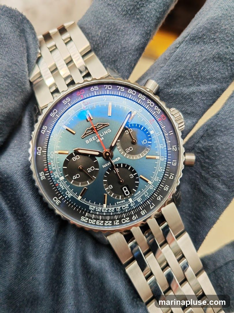 A close-up shot of a Breitling Navitimer watch with a blue dial and silver metal band. The watch is held by a gloved hand. The dial features sub-dials and a slide rule bezel.