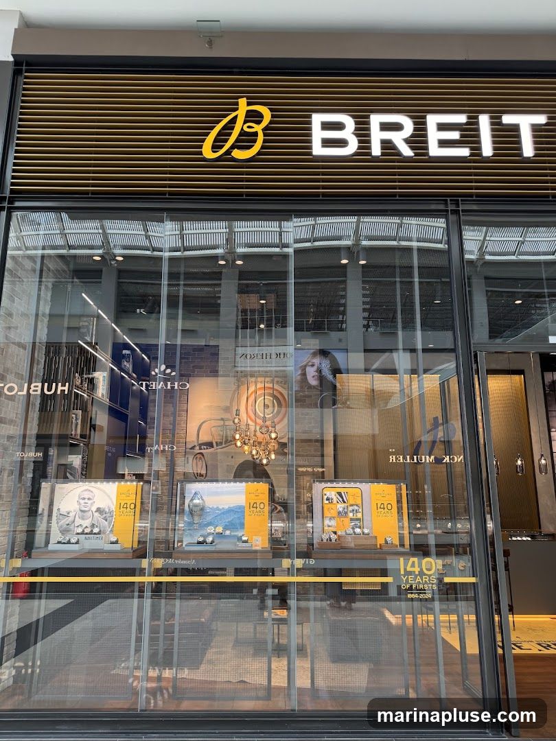 The storefront of a Breitling watch store. The store has a large glass facade with the Breitling logo and name prominently displayed above the entrance. Inside, display cases showcase watches and promotional materials celebrating "140 Years of Firsts 1884-2024". Other watch brands like Hublot and Franck Muller are also visible in reflections or adjacent displays.