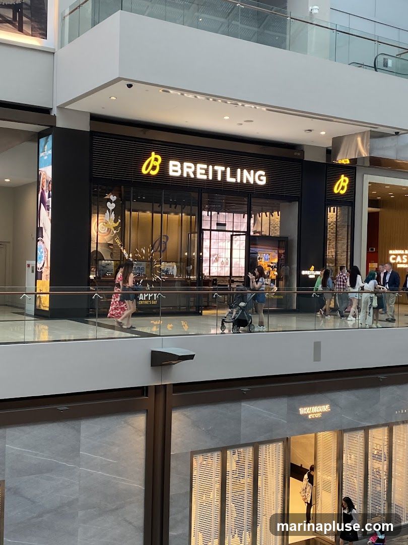 A Breitling watch store is visible from a higher level in a shopping mall. The store has a prominent illuminated sign that reads "BREITLING" with the brand's logo. The storefront features large glass windows displaying watches and interior lighting. People are seen walking past the store on the level above.
