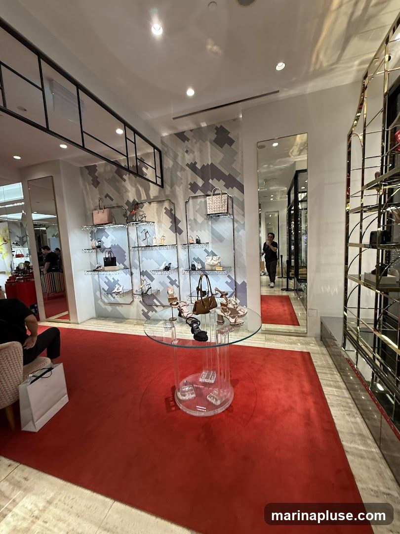 The interior of a high-end retail store, likely a shoe and handbag boutique. A plush red carpet covers the floor, leading to display shelves filled with various women's shoes and handbags. A circular glass table in the foreground showcases more footwear. In the background, a person is visible walking through a doorway, and mirrors reflect the store's elegant design. The walls feature a modern, geometric pattern.
