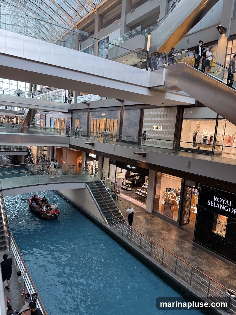 A view of a modern indoor shopping mall with a canal running through it. A boat with passengers navigates the blue water. Multiple levels of shops and escalators are visible, with people browsing and walking. Brands like Chloe, Pazzion, and Royal Selangor are identifiable.