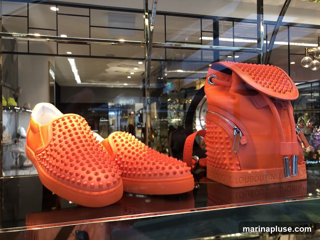 A display window showcases a pair of bright orange slip-on shoes and a matching backpack, both adorned with the brand's signature spikes. The shoes are positioned in the foreground, with one slightly ahead of the other. The backpack is to the right, standing upright. The items are displayed on a reflective surface, and the background shows a blurred interior of a store with shelves of other merchandise and lighting fixtures. The brand name 'LOUBOUTIN' is visible embossed on the base of the backpack.