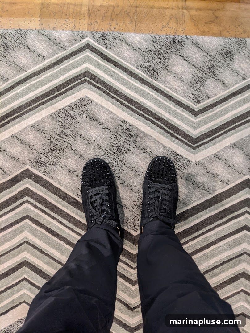 A top-down view of a person wearing black studded sneakers and dark pants, standing on a chevron-patterned rug. The rug has alternating bands of grey, white, and dark grey. The floorboards are visible at the top of the image.
