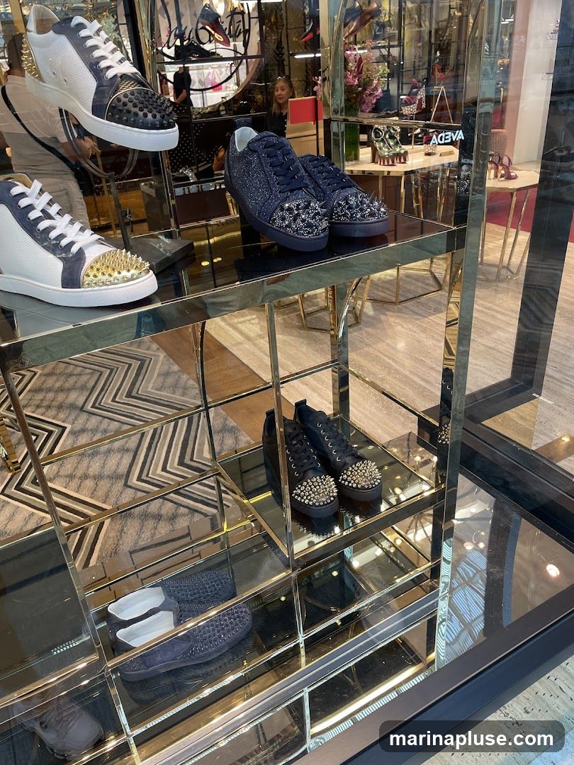 A display of luxury footwear, including sneakers with spikes and glitter, and loafers, is showcased on mirrored shelves in a store window. The background shows a glimpse of the store's interior with more shoes and decorative elements.