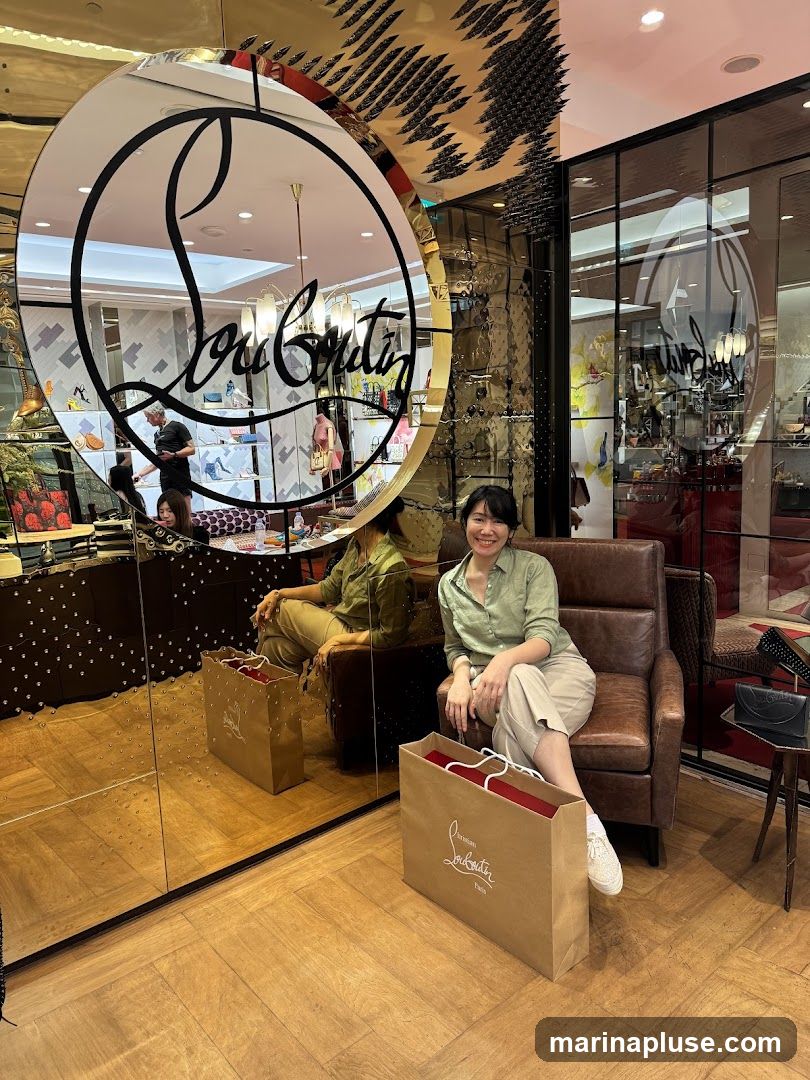 A woman sits in a leather armchair in a brightly lit store, with a large shopping bag from Christian Louboutin at her feet. The store's interior is reflected in a large, ornate mirror, showing displays of shoes and handbags. The woman is wearing a light green collared shirt and beige pants.