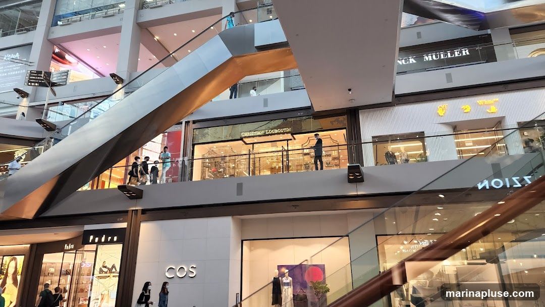 The image shows the interior of a modern shopping mall with multiple levels and escalators. Several storefronts are visible, including Christian Louboutin, Off-White, and COS. People are seen walking on escalators and browsing in the shops. The architecture is sleek and contemporary, with glass railings and metallic accents.