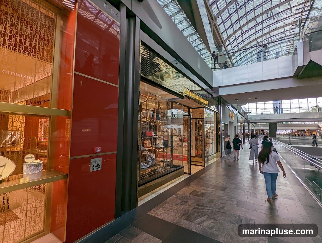 A view of several high-end retail stores within a modern shopping mall. The storefronts feature large glass windows displaying merchandise, with one store prominently featuring the name "CHRISTIAN LOUBOUTIN". The architecture includes a distinctive glass ceiling that allows natural light to filter in. People are seen walking along the polished floor of the mall, with escalators and upper levels visible in the background.