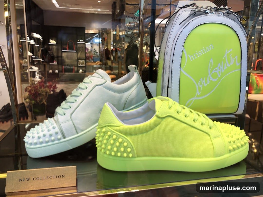 A display window featuring a neon yellow Christian Louboutin backpack and two pairs of low-top sneakers, one in mint green and the other in neon yellow. The sneakers have the signature Louboutin spike detailing on the toe. A small sign in the foreground reads "NEW COLLECTION". The background shows a blurred interior of a luxury store with shelves of shoes and handbags.