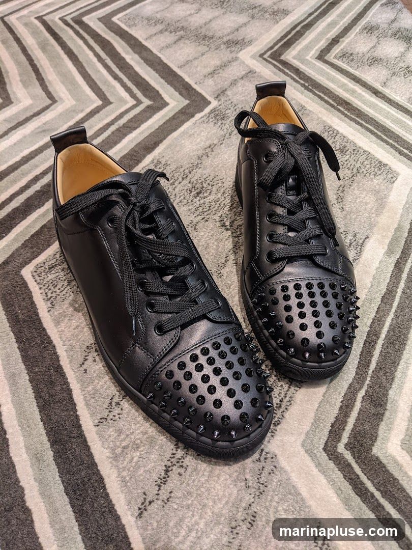 A pair of black leather low-top sneakers with black laces and black studs on the toe box. The sneakers are placed on a rug with a geometric pattern.