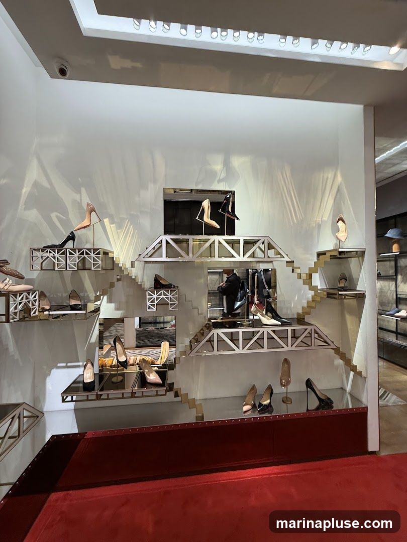 A well-lit display of various high-heeled shoes arranged on tiered shelving units that resemble staircases and bridges. The shoes are presented in a modern and elegant fashion, with some on individual stands and others placed directly on the shelves. The background is white, with subtle lighting creating shadows and highlights. A red carpet runs along the bottom of the display.