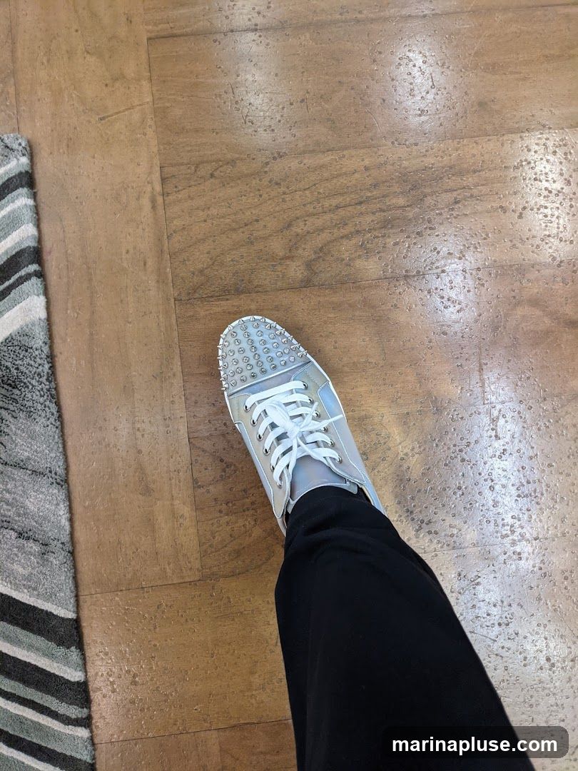 A person is wearing silver sneakers with white laces and silver spikes on the toe. They are also wearing black pants. The sneakers are on a wooden floor next to a striped rug.