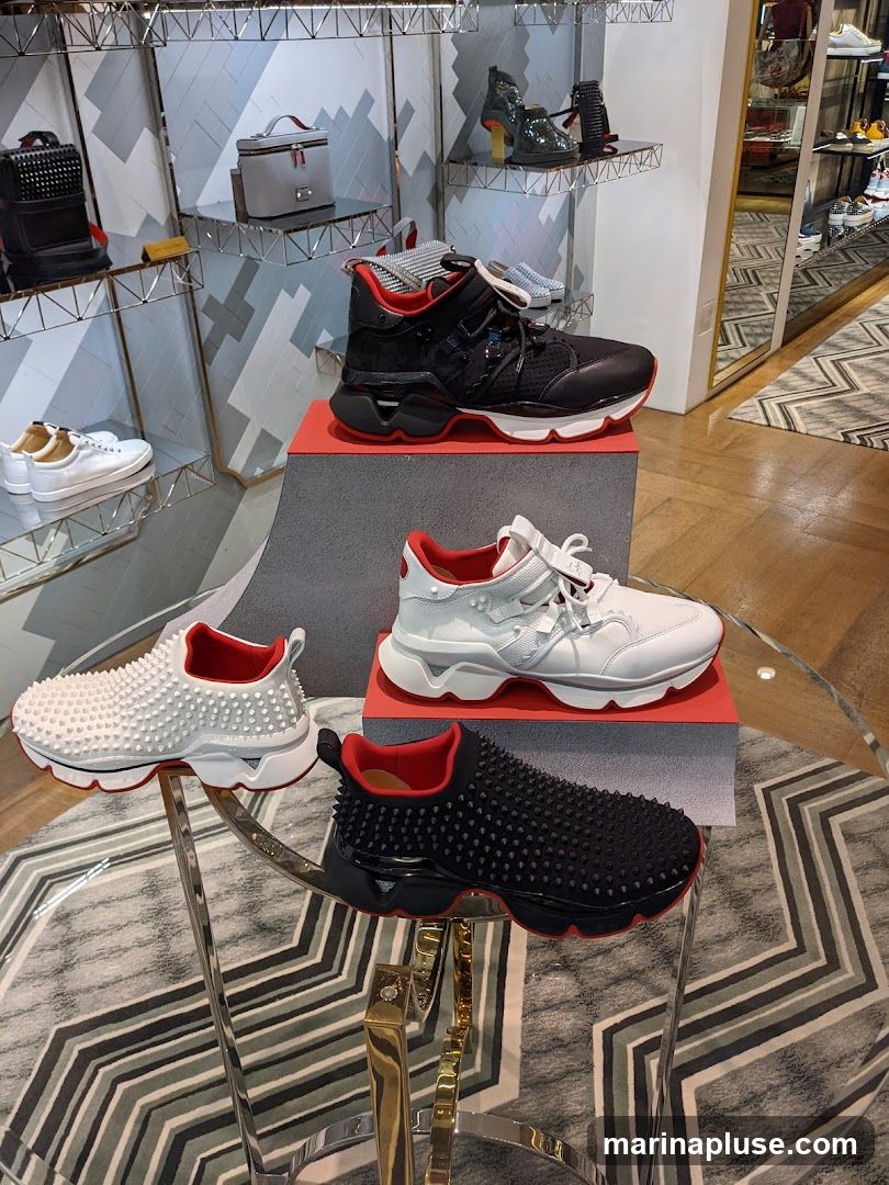 A display of designer sneakers and handbags in a retail store. The sneakers are showcased on tiered stands and a glass table, with various styles and colors visible. In the background, shelves hold handbags and other footwear, and a mirrored wall reflects the store's interior.