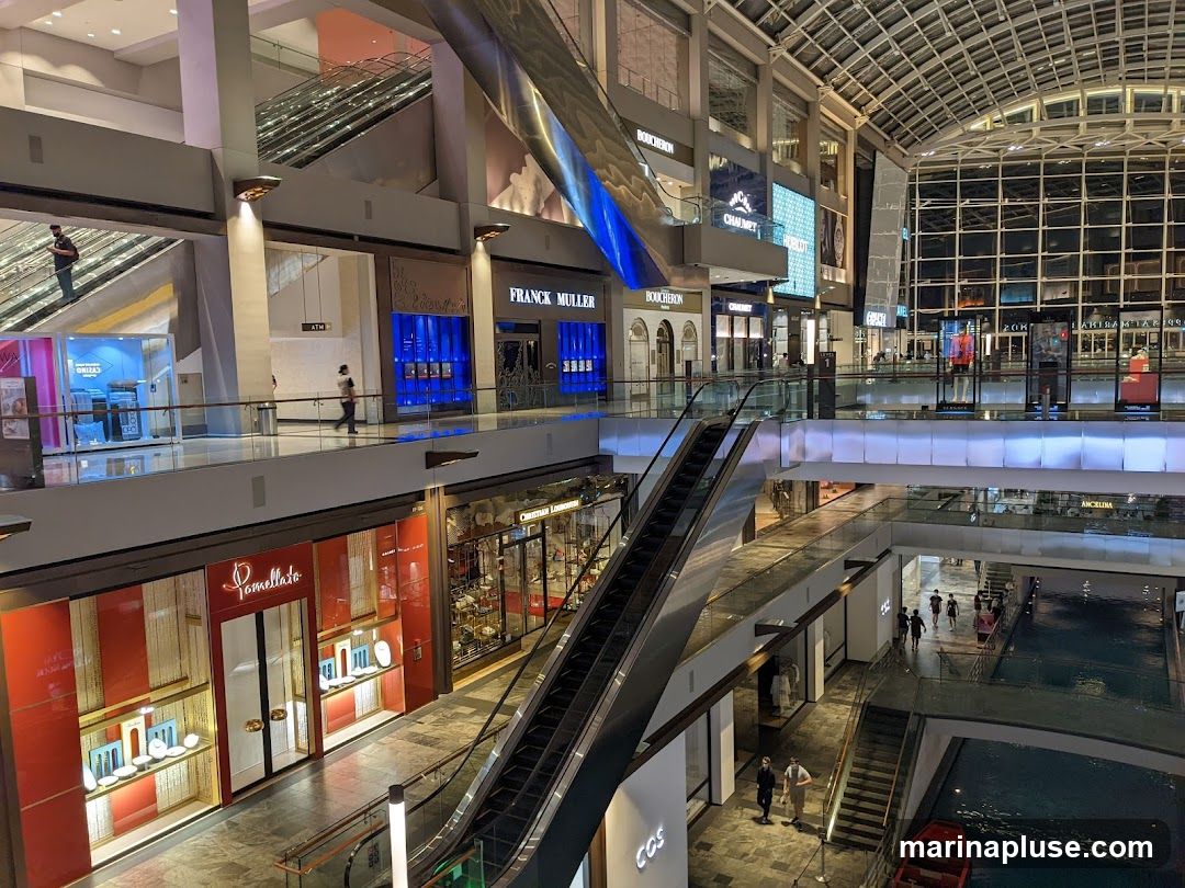 The image shows the interior of a large, modern shopping mall. Escalators connect different levels, and various shops are visible along the walkways. Some shops display luxury goods, such as jewelry and watches, with illuminated displays. A canal or water feature is visible on the lower level, with a small red boat. People are seen walking around the mall. The architecture features a high, glass-paneled ceiling allowing natural light.