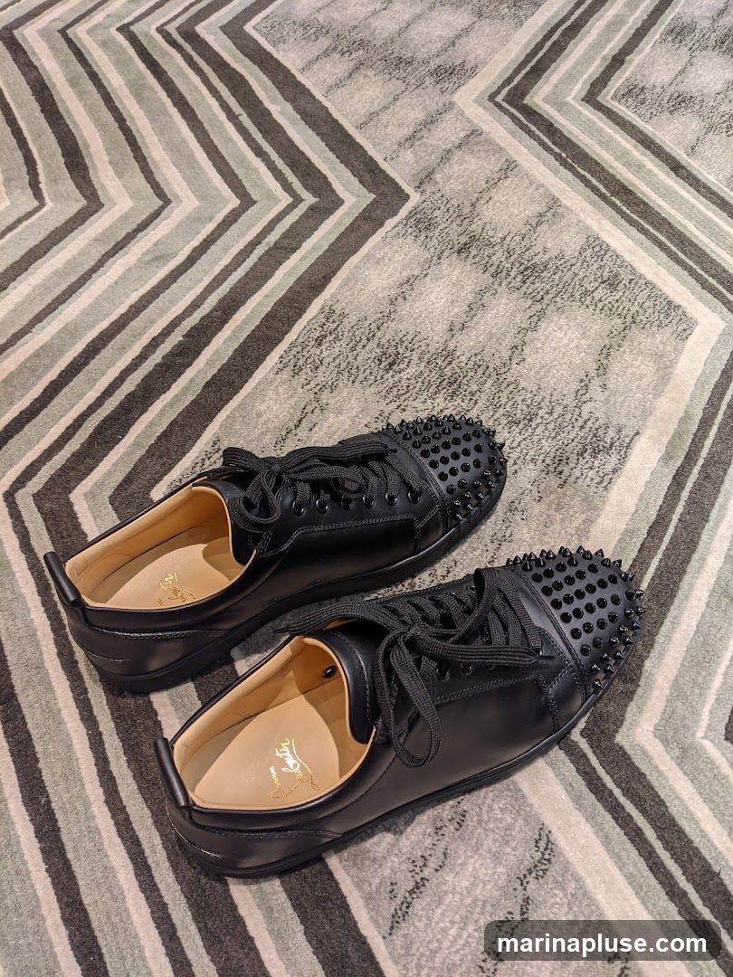 A pair of black leather low-top sneakers with black spikes on the toe box. The shoes are placed on a rug with a geometric pattern of grey and white stripes. The insoles of the shoes have a gold logo that appears to be Christian Louboutin.