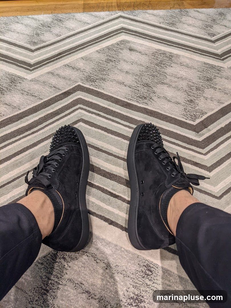 A pair of black suede sneakers with black spikes on the toe box and black laces. The shoes are worn by a person wearing black pants and no-show socks. The sneakers are on a rug with a chevron pattern of grey and white stripes.