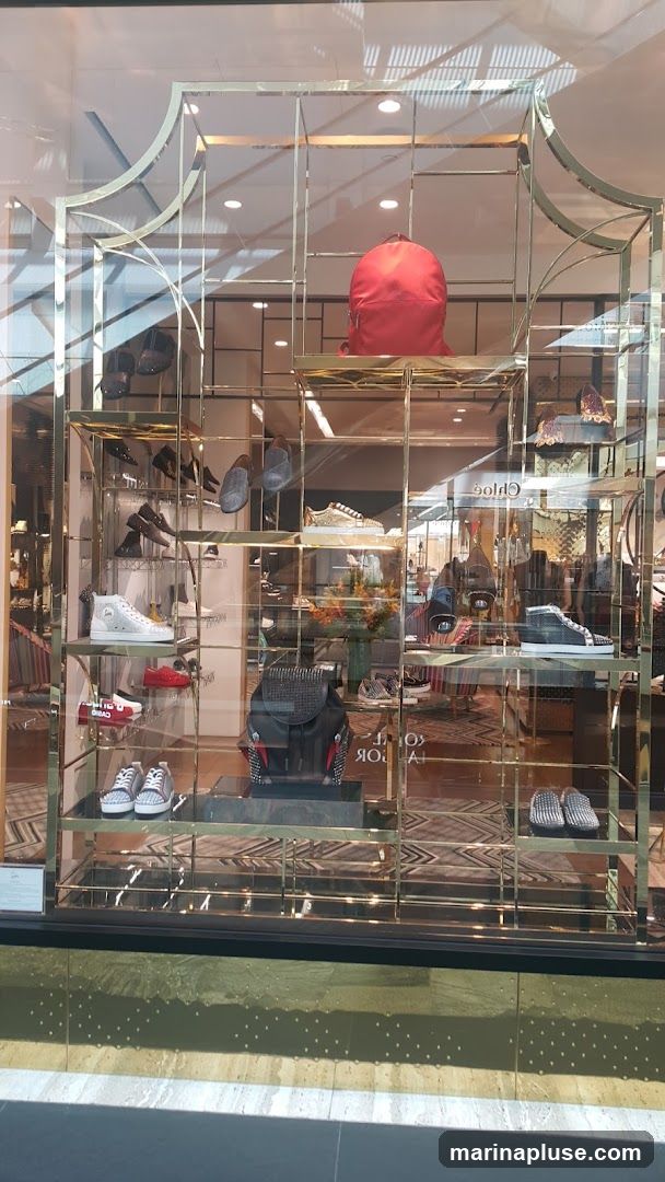 A store window display featuring various luxury accessories, including handbags, shoes, and a red backpack. The items are arranged on gold shelving units. The reflection of the interior and the exterior can be seen on the glass.