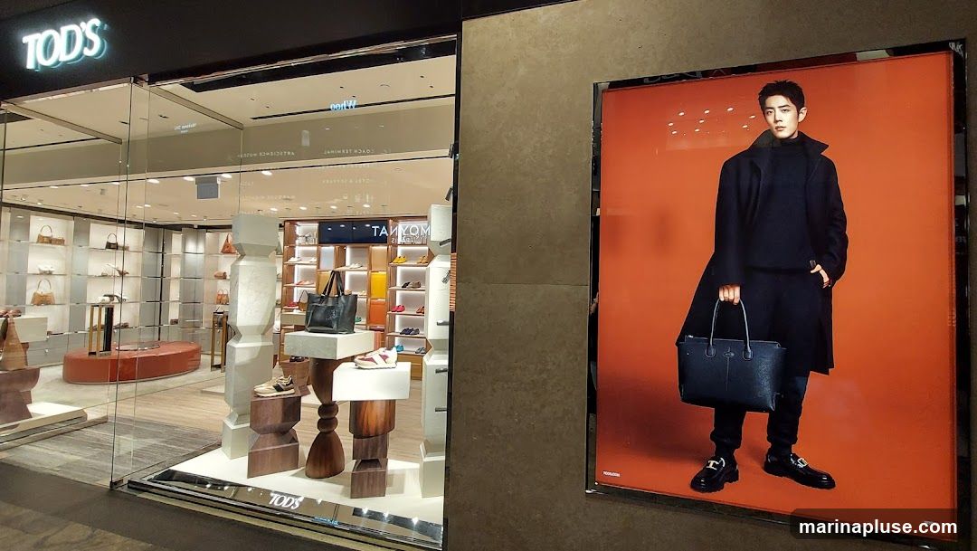 The image shows the exterior of a TOD'S store with its name visible on the facade. Inside, shelves display handbags and shoes. To the right, a large advertisement features a man in a dark coat and pants, holding a black handbag and wearing black loafers. The advertisement has an orange background.