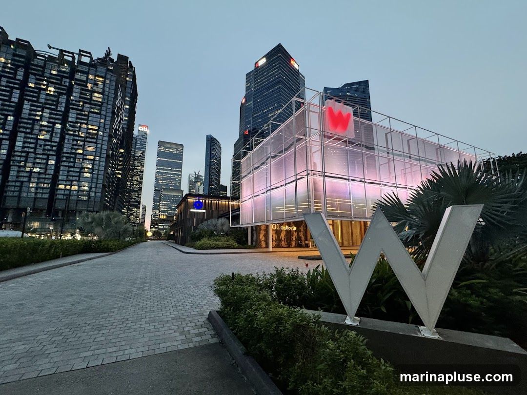 A modern building with a large W logo in front, illuminated by pink and purple lights. The building has a glass facade and is surrounded by lush greenery and palm trees. In the background, several tall skyscrapers are visible against the twilight sky.