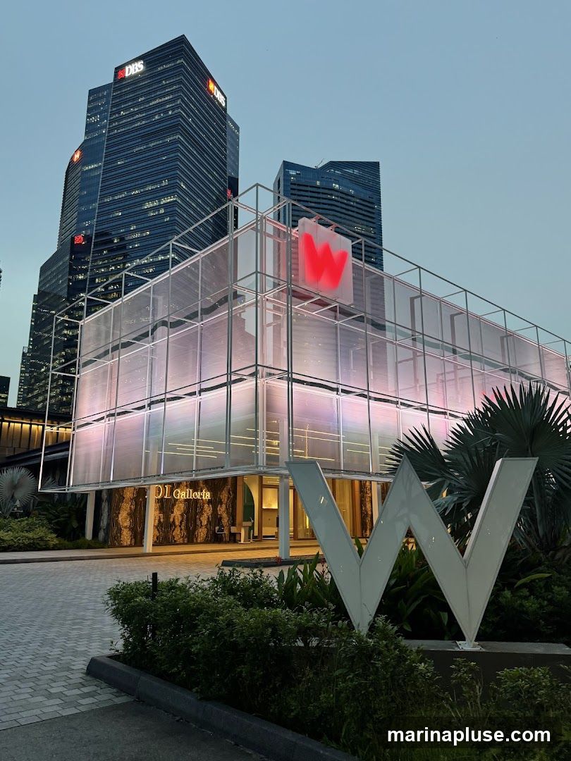 The exterior of a modern building with a large illuminated 'W' logo in front. The building features a glass facade with colorful lighting and a prominent sign that reads 'DI Galleria'. Tall skyscrapers are visible in the background.