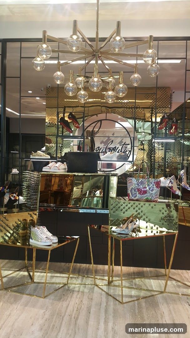 A well-lit display inside a luxury shoe store. The display features a variety of high-end footwear, including sneakers and heels, presented on gold-colored stands and shelves. A large circular mirror with a prominent logo is visible in the background. The store's interior is sleek and modern, with mirrored surfaces and a sophisticated chandelier.