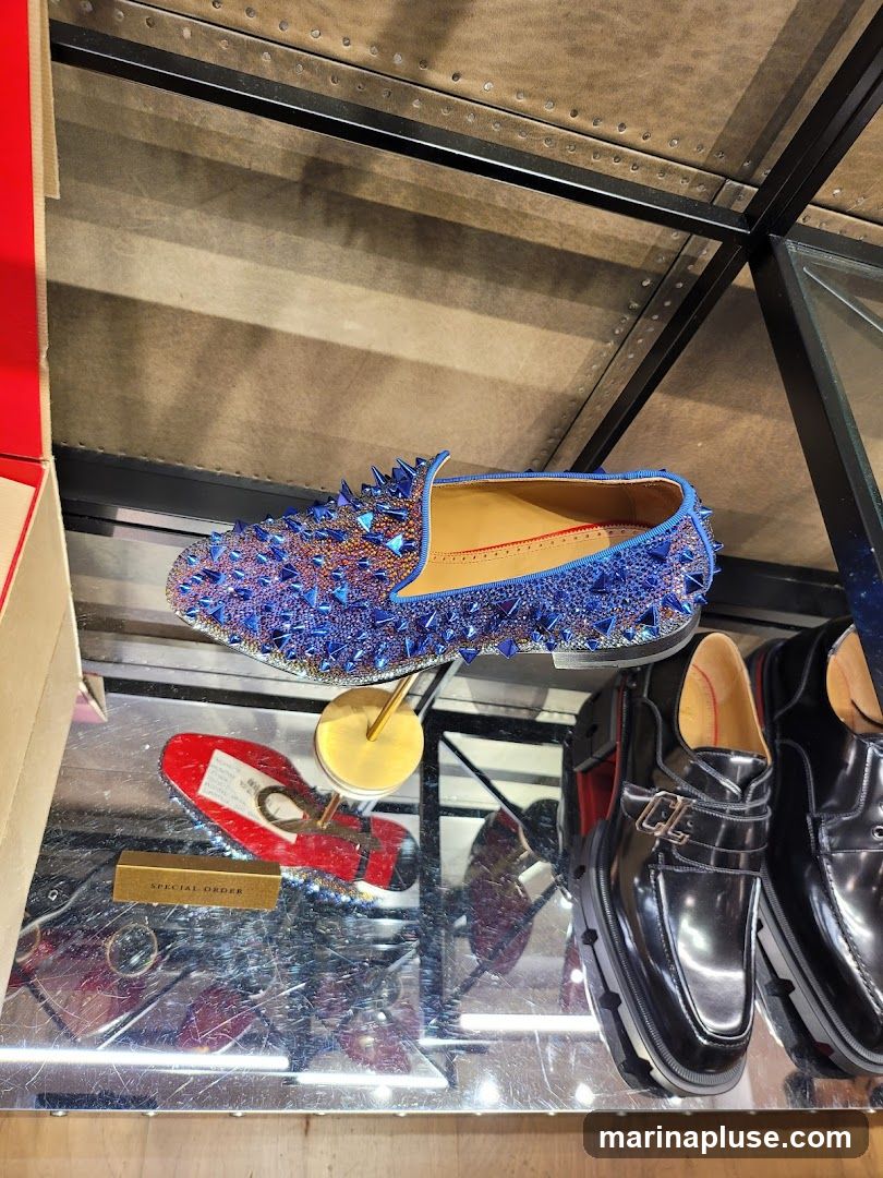 A display of designer shoes, including a blue loafer covered in spikes and rhinestones, and a pair of black patent leather loafers with buckle detailing. A red shoe with a white tag is also visible, along with a small gold box labeled "SPECIAL ORDER". The shoes are displayed on a reflective surface under shelving.