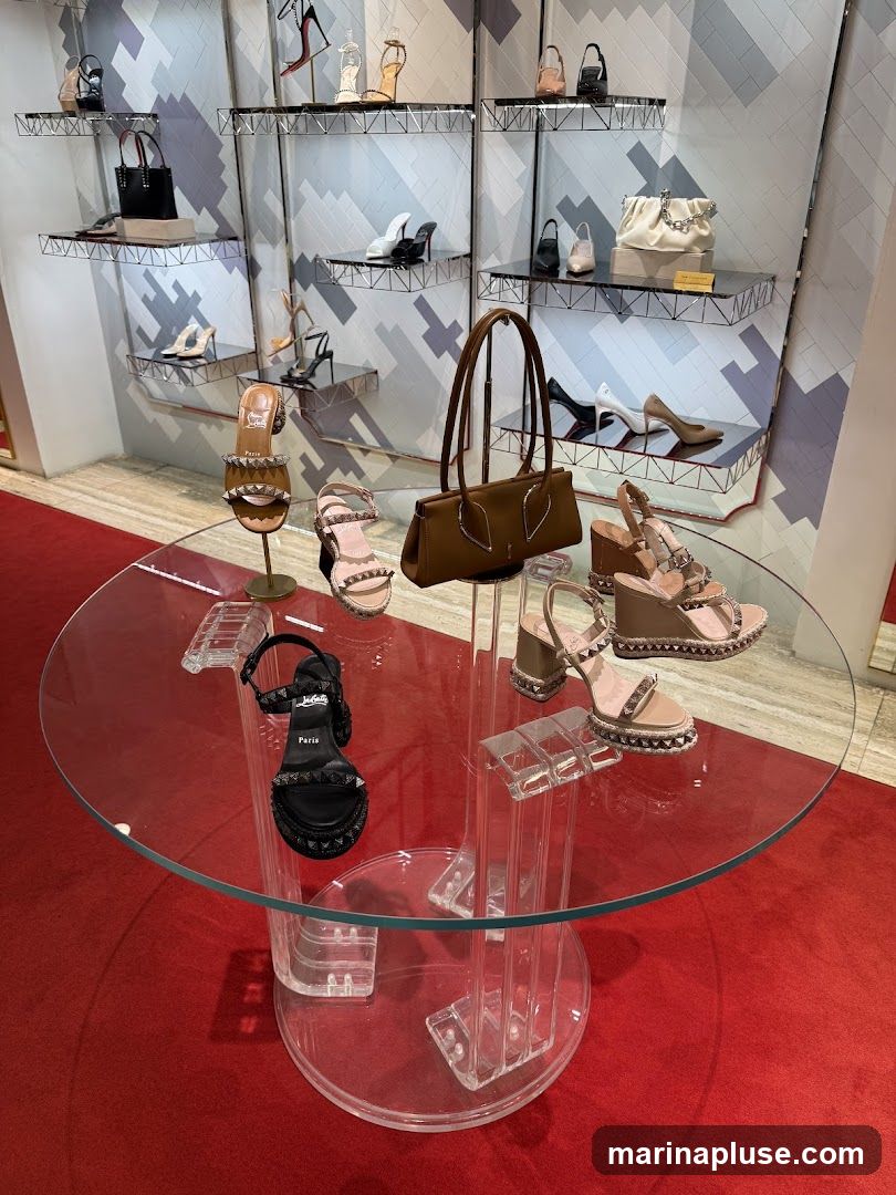 A display of high-end women's footwear and handbags is showcased on tiered shelving and a glass table. The collection features various styles of heels, sandals, and wedges, many adorned with studs and embellishments. Several designer handbags are also visible, including a brown tote and a cream-colored clutch. The display is set against a modern, geometric-patterned wall and a plush red carpet.