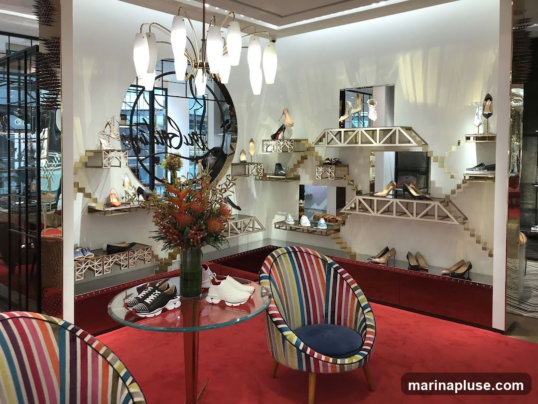 A brightly lit interior of a shoe store with a prominent circular mirror displaying a signature logo. Various styles of high-heeled shoes and sneakers are displayed on tiered, geometric shelving units. A glass table in the foreground holds a pair of black studded sneakers and white studded sneakers. Two colorful striped armchairs are placed on a red carpet. A vase of orange and red flowers adds a decorative touch.