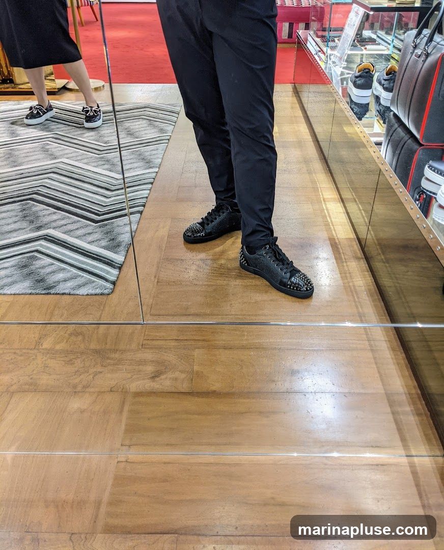 A person is standing in a store, wearing black pants and black sneakers with silver spikes on the toe. In the reflection of a mirror, another person is visible wearing black shoes with white bows. The store appears to be a high-end boutique, with displays of handbags and other luxury items visible in the background.