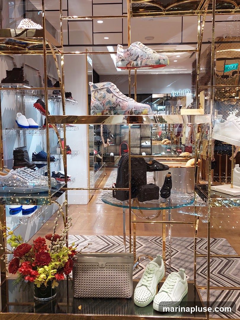 A display of high-end footwear and accessories in a store window. The shelves showcase various sneakers and high-top shoes with intricate designs and embellishments, including studded details and floral patterns. A clutch bag and a bouquet of flowers are also visible. In the background, the store interior features more merchandise and signage, including a visible "Chloe" logo.