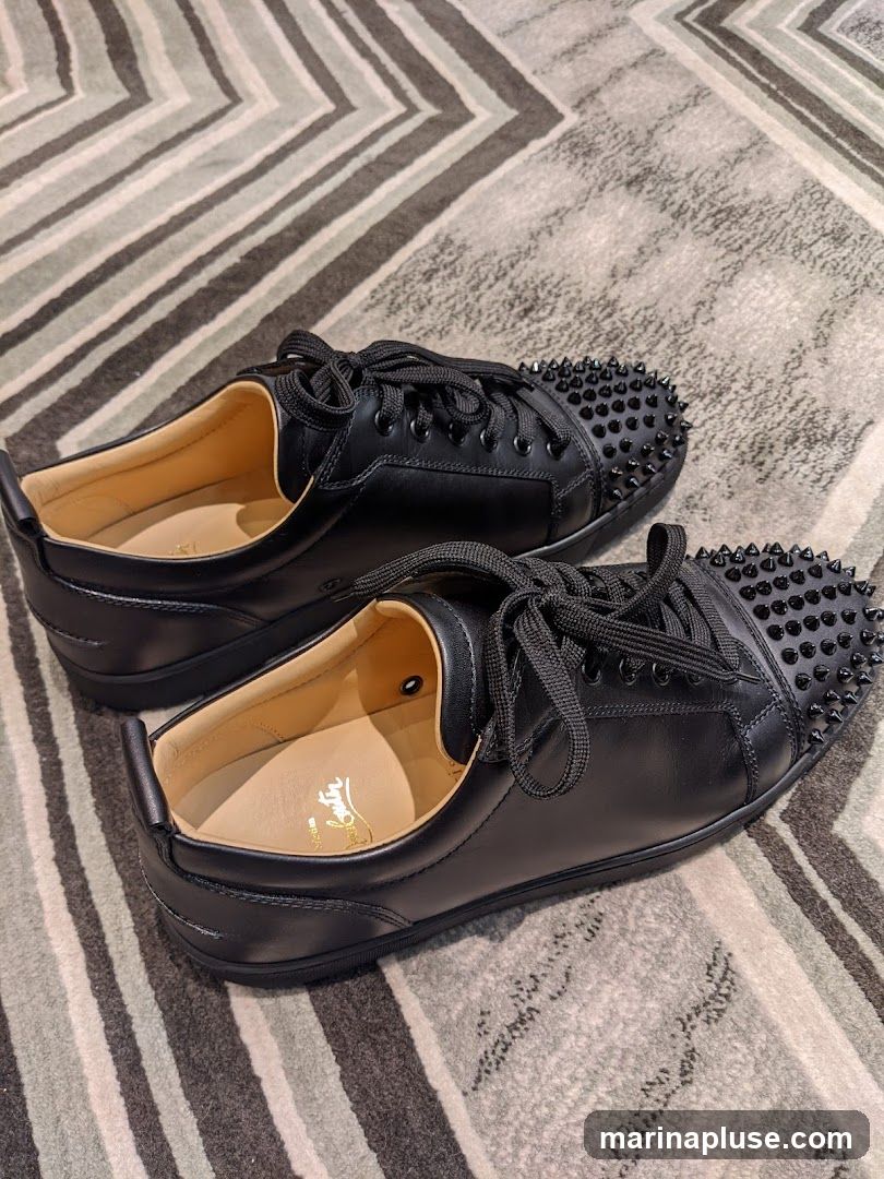 A pair of black leather Christian Louboutin sneakers with black spikes on the toe box. The shoes are on a striped carpet. The Louboutin logo is visible on the insole of the left shoe.