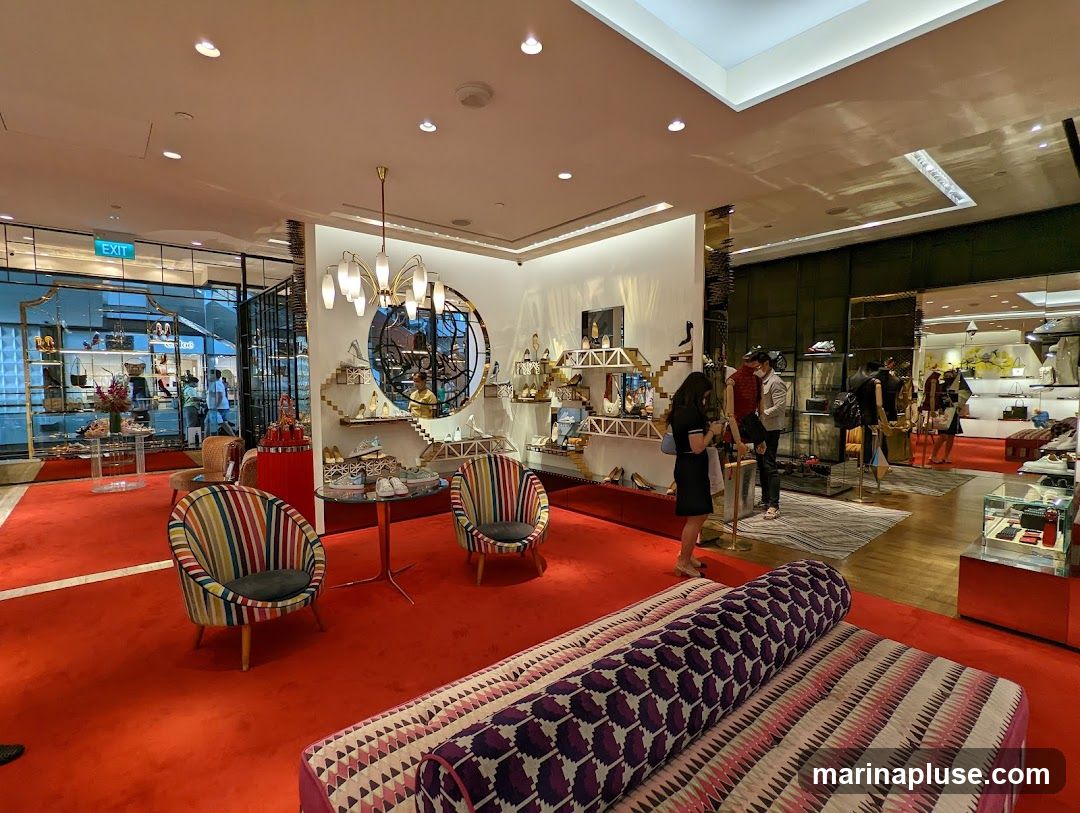 The interior of a high-end shoe store with a vibrant red carpet. Display shelves showcase various shoes and handbags. Several customers are browsing the merchandise, and staff members are assisting them. The store features modern lighting fixtures and decorative elements, including colorful striped armchairs and a patterned sofa.