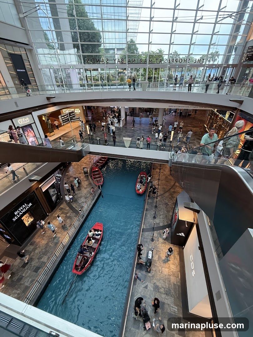 An interior view of a large, multi-level shopping mall with a canal running through the center. Red gondola-style boats navigate the blue water. People are seen shopping, walking, and riding escalators on various levels. Storefronts for brands like Chloe and Royal Selangor are visible. The mall features a glass ceiling and modern architecture.