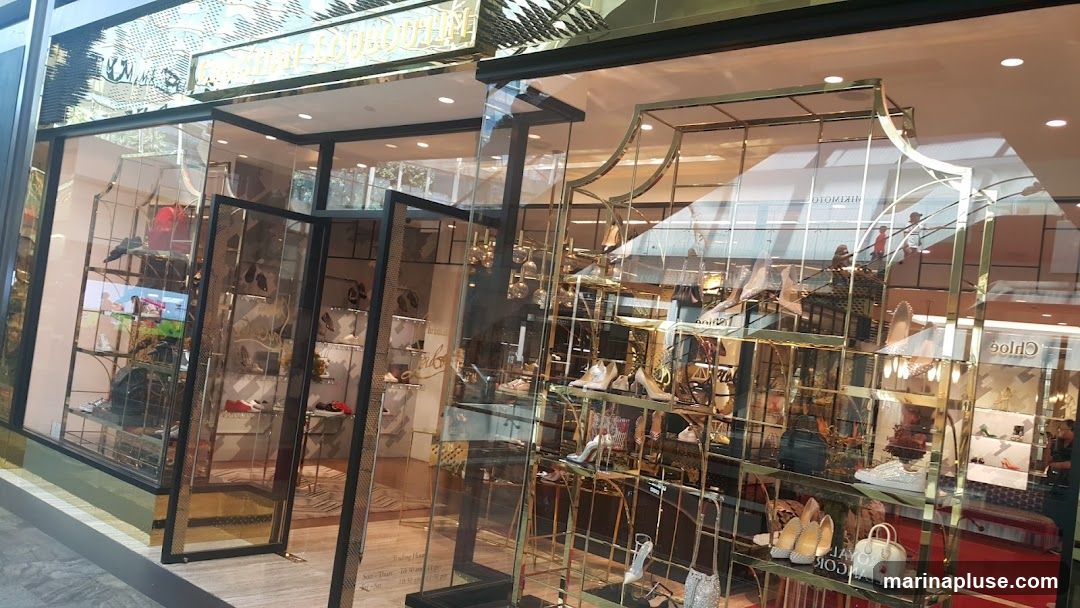 The exterior of a Christian Louboutin shoe store. The storefront features large glass windows displaying various high-heeled shoes, sneakers, and handbags on gold shelving units. The store's name is visible in gold lettering above the entrance. Reflections of people and escalators are visible in the glass.