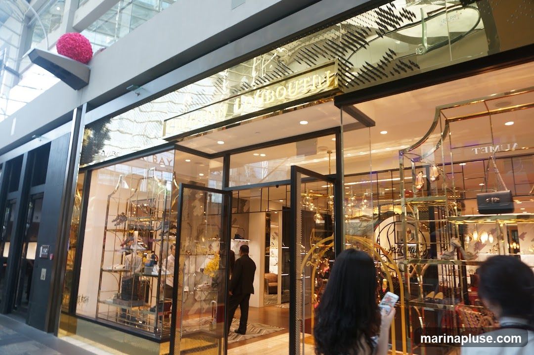 A wide shot of a luxury fashion boutique's storefront. The entrance features double glass doors, and the facade is adorned with gold accents and intricate patterns. Display windows showcase various handbags and shoes. The store's name, partially visible as "...BAY" and "...BOUTIQU", is displayed in prominent gold lettering above the entrance. Inside, mannequins and shelves display merchandise.
