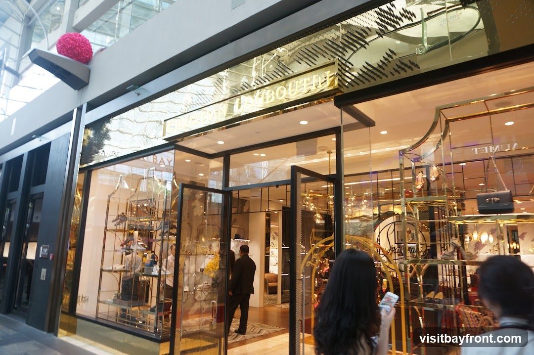 A wide shot of a luxury fashion boutique's storefront. The entrance features double glass doors, and the facade is adorned with gold accents and intricate patterns. Display windows showcase various handbags and shoes. The store's name, partially visible as "...BAY" and "...BOUTIQU", is displayed in prominent gold lettering above the entrance. Inside, mannequins and shelves display merchandise.
