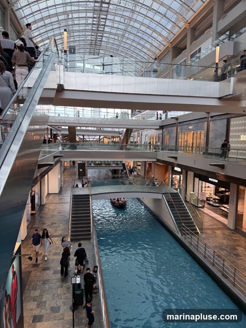An interior view of a multi-level shopping mall with a canal running through it. People are seen walking on escalators, stairs, and the ground floor. A boat is navigating the canal. Shops are visible on various levels, with some displaying merchandise. The architecture features a large, arched glass ceiling.