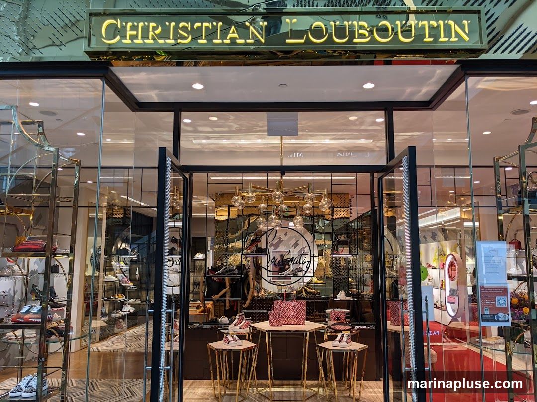 The exterior of a Christian Louboutin store. The store has large glass windows and doors, with the brand name "CHRISTIAN LOUBOUTIN" displayed in gold lettering above the entrance. Inside, shelves are stocked with shoes and bags.