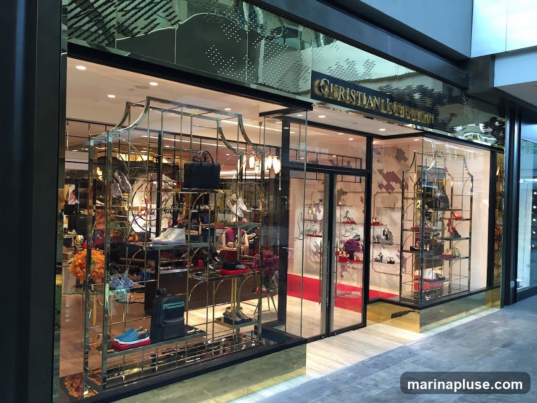 The exterior of a Christian Louboutin store, featuring a large display window showcasing various shoes and handbags. The store's name is prominently displayed in gold lettering above the entrance. The interior is visible through the glass, with shelves displaying merchandise and a red carpet leading into the store.