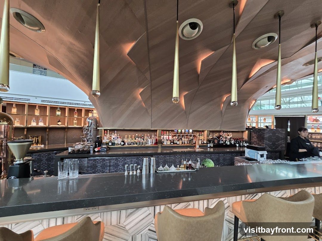 A view of a modern bar interior with a long counter, stools, and a well-stocked back bar. The ceiling features a unique, curved wooden design with pendant lights. Bottles of alcohol are neatly arranged on shelves behind the bar, and a barista is visible on the right.