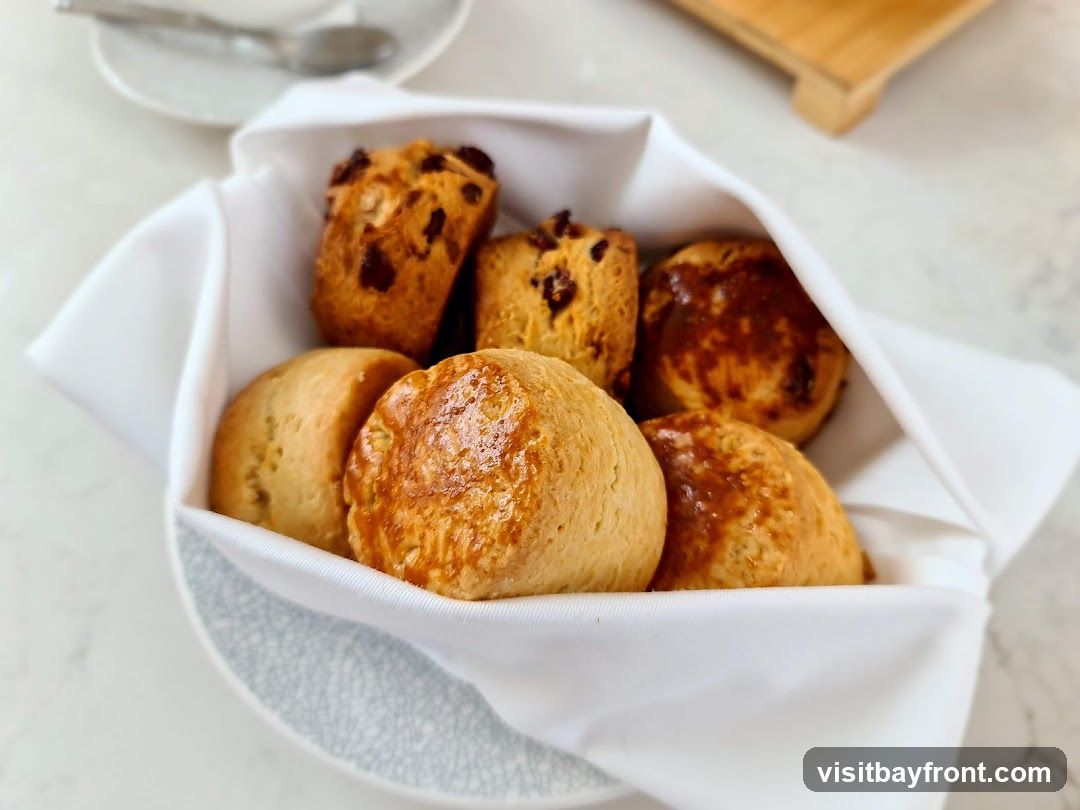 A white cloth-lined basket holds an assortment of scones. Some scones are plain and golden brown, while others appear to have dried fruit or chocolate chips baked into them. The basket sits on a light-colored surface, possibly a marble countertop, with a blurred background showing a teacup and saucer.