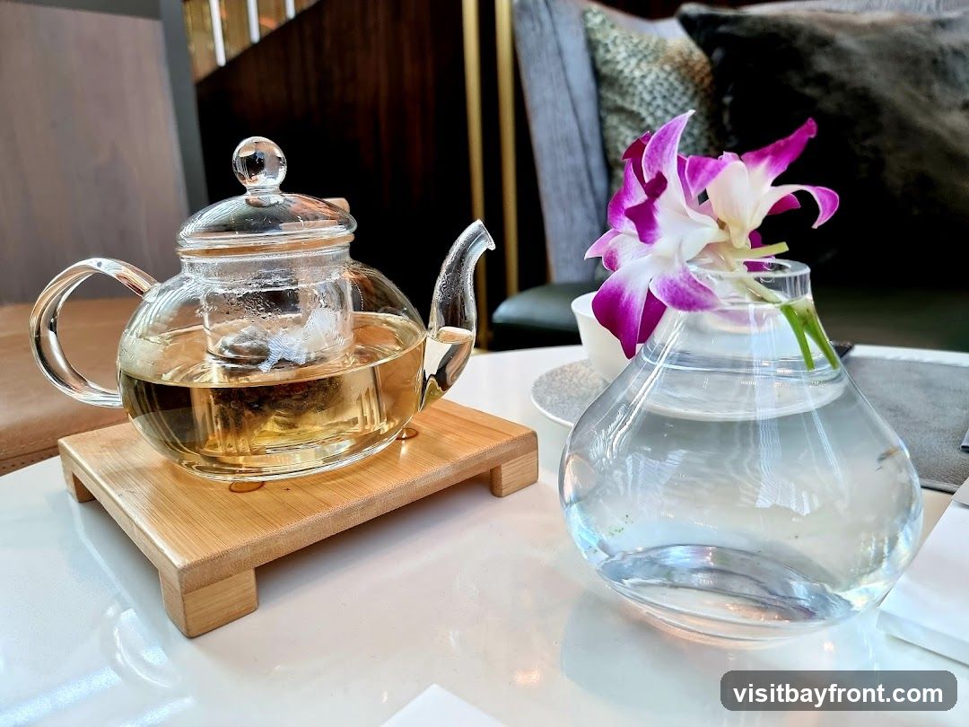 A clear glass teapot filled with amber-colored tea sits on a bamboo stand. Next to it, a small glass vase holds a single orchid with purple and white petals. The setting appears to be indoors, possibly a lounge or dining area, with soft lighting and comfortable seating visible in the background.