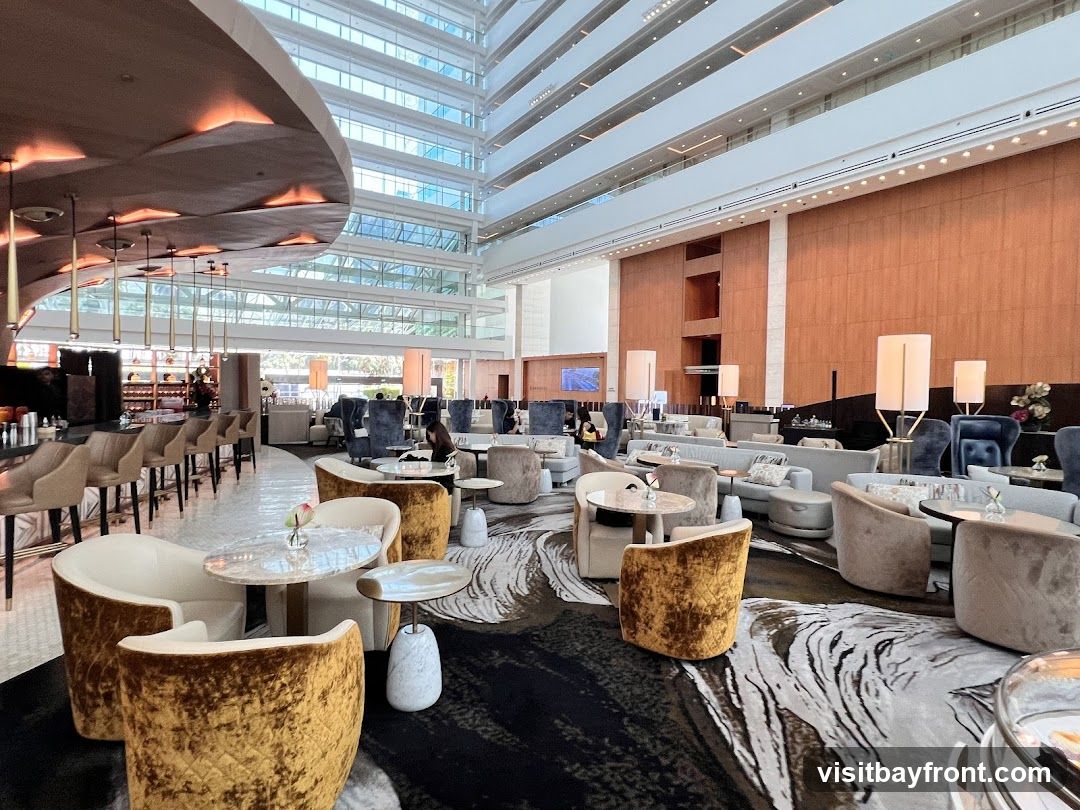 A spacious and elegantly designed lounge area with a bar, comfortable seating, and a multi-story atrium. The decor features plush velvet chairs in gold and grey, marble-topped tables, and a striking patterned carpet. Tall lamps and modern light fixtures illuminate the space, which is set against a backdrop of wood-paneled walls and a glass-enclosed atrium. Several patrons are seated, enjoying the ambiance.