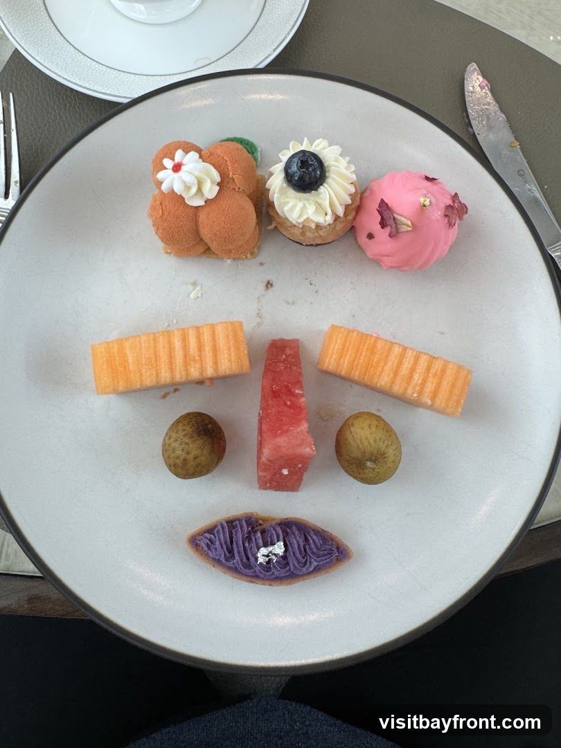 A white plate holds a dessert platter arranged to resemble a face. The eyes are represented by two whole longans. The nose is a slice of watermelon. The mouth is a purple pastry with silver leaf. The forehead is adorned with three small desserts: one orange-colored with white frosting and a red flower, one with white frosting and a blueberry, and a pink spherical dessert with gold leaf and rose petals. Sliced cantaloupe forms the cheeks. A fork and knife are visible on the table.