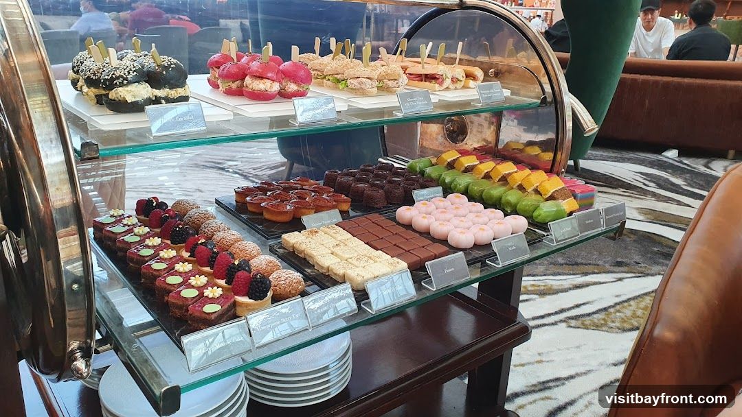 A display of various desserts and small sandwiches on tiered shelves. The desserts include tarts, cakes, pastries, and colorful mochi. Small sandwiches and sliders are also visible.