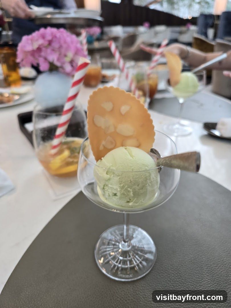 A close-up shot of a dessert featuring two scoops of light green ice cream in a stemmed glass, topped with a thin, round cookie adorned with almond slivers. The dessert is placed on a dark gray placemat. In the blurred background, there are other drinks and floral arrangements, suggesting a dining setting.