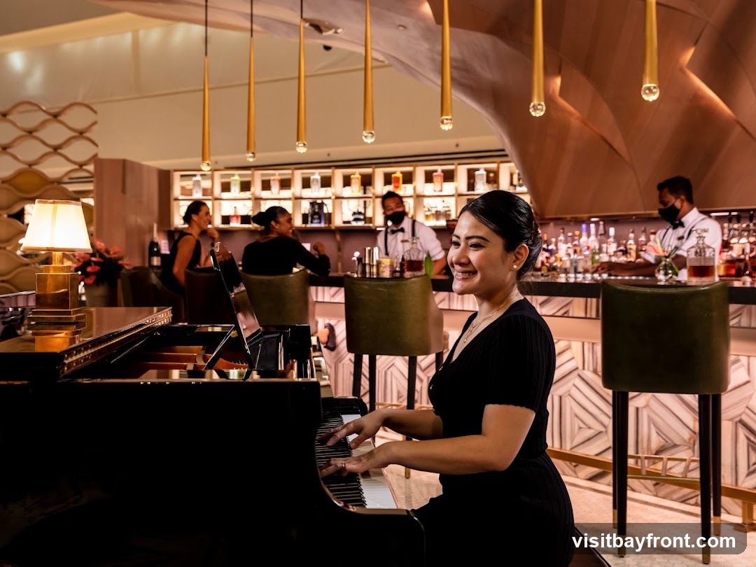 A woman in a black dress plays a grand piano in a dimly lit bar. Behind her, bartenders in masks serve drinks at a well-stocked bar. Several patrons are seated at the bar, and decorative pendant lights hang from the ceiling.