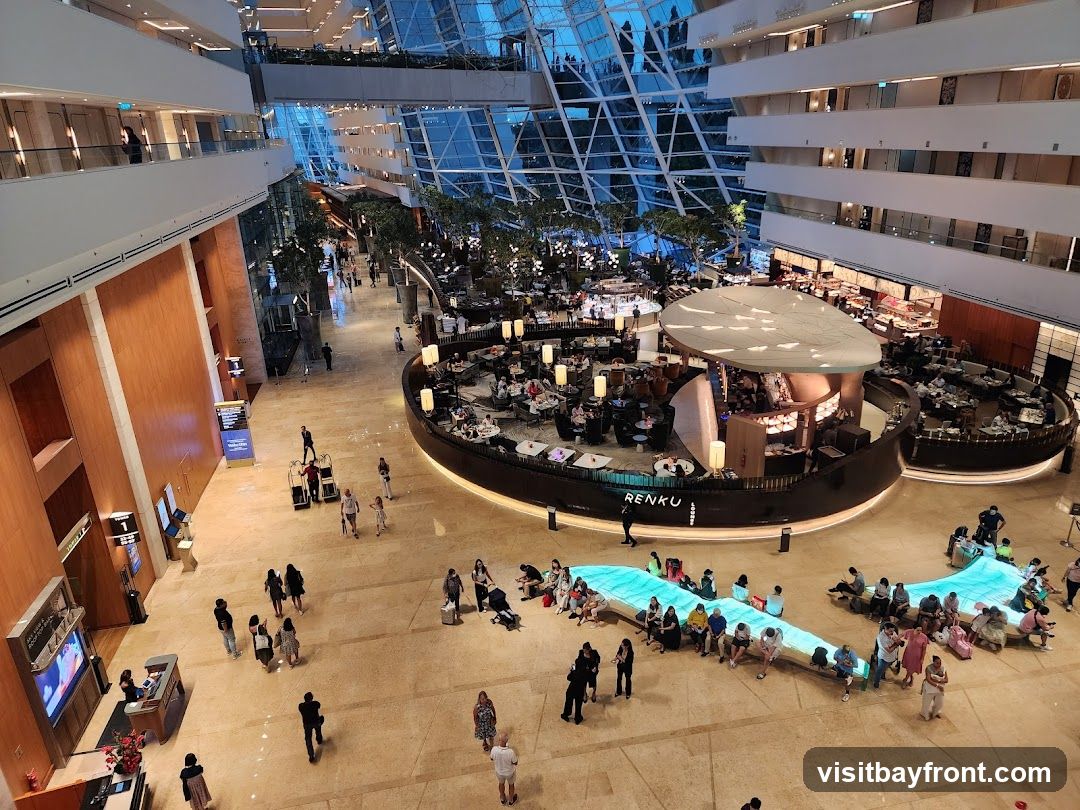 An aerial view of a spacious, multi-level atrium with a modern architectural design. The ceiling features a geometric glass structure, allowing natural light to filter in. In the center, a circular, elevated dining area with a distinctive roof is surrounded by tables and chairs, with people dining. To the right, another dining area is visible. In the foreground, a long, illuminated bench-like structure is occupied by a group of people. The floor is polished marble, and there are various plants and trees throughout the space. People are seen walking and sitting in different areas of the atrium. A sign with the word "RENKU" is visible on the central dining structure.